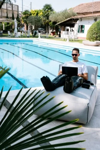 Uomo seduto su un lettino a bordo piscina mentre utilizza il computer portatile e indossa i gambali di pressoterapia Wepere VIGGO.